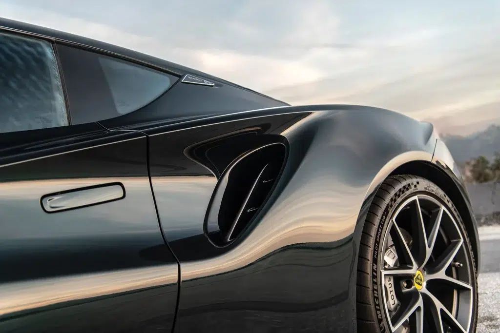 Seitenansicht eines dunklen Lotus Emira Sportwagens mit Fokus auf Hinterrad, Lufteinlass und glänzender Karosserie.