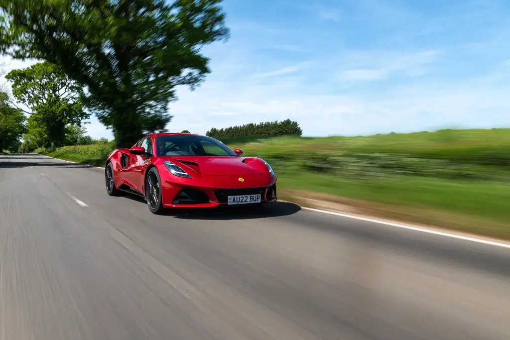 Roter Lotus Emira Sportwagen fährt mit Bewegungsunschärfe auf einer Landstraße durch grüne Landschaft unter blauem Himmel.