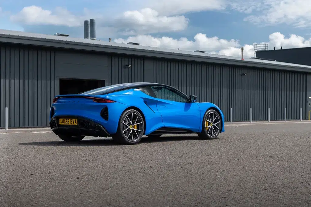 Blauer Lotus Emira Sportwagen in Heckansicht vor einem grauen Industriegebäude.