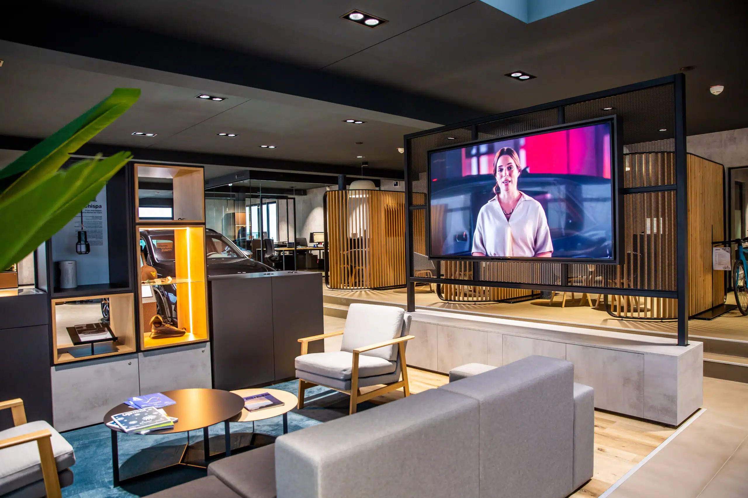 Moderner Showroom-Lounge mit großem Bildschirm, der eine Frau zeigt, Sitzbereichen und dahinterliegendem Büro.