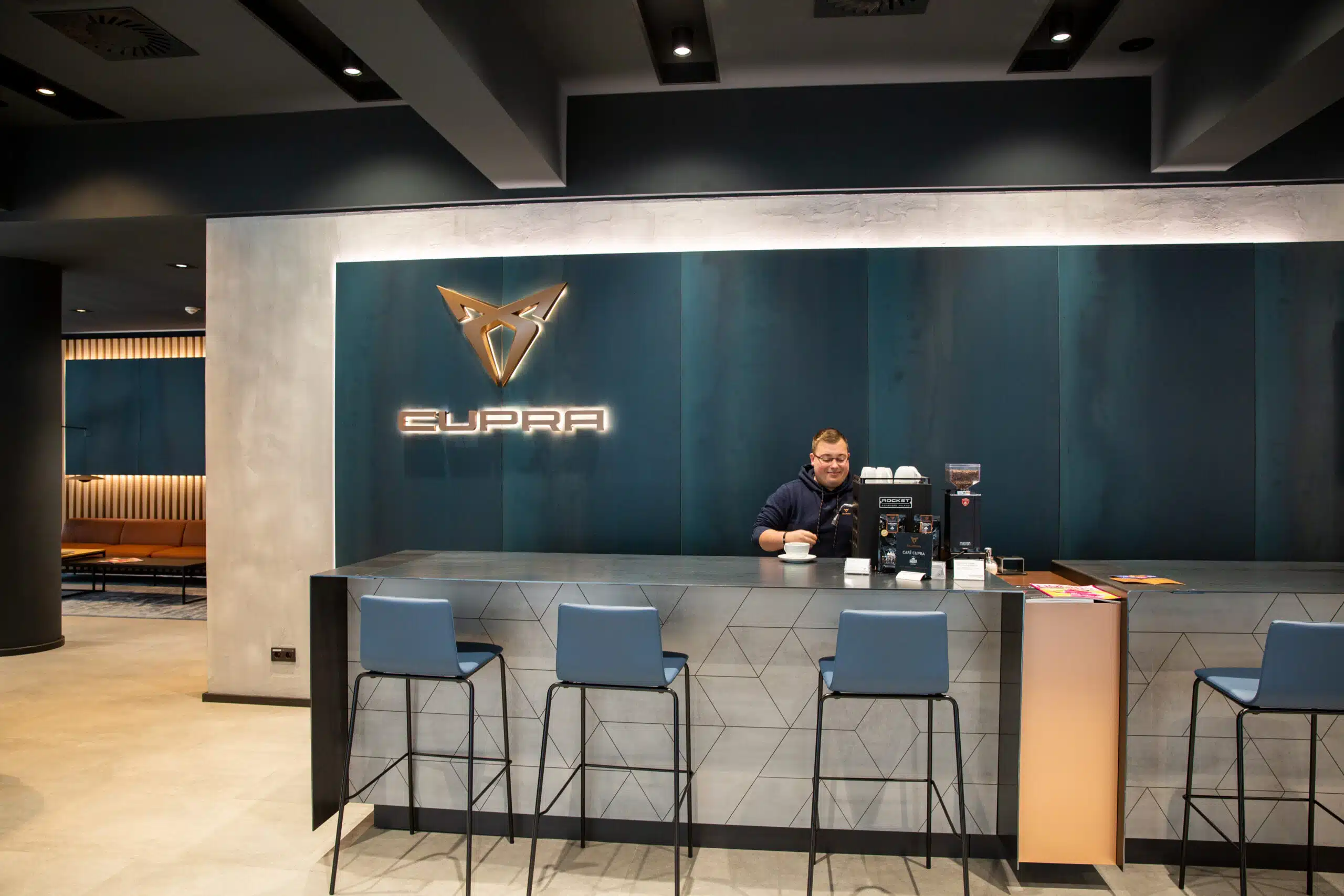 Ein Mann bereitet Kaffee an einer modernen Theke mit Cupra-Logo im Hintergrund zu.