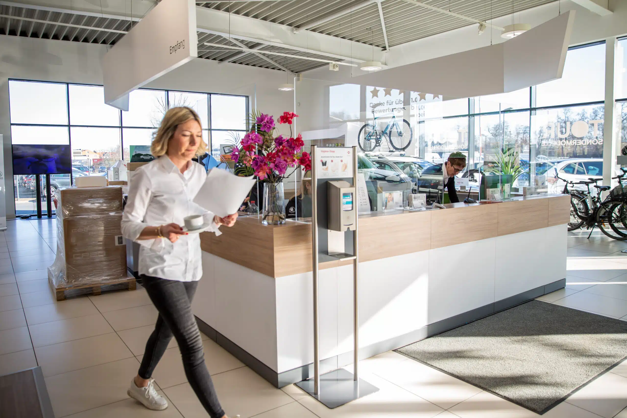 Eine Frau mit Papieren und einer Tasse geht durch den hellen Empfangsbereich eines Autohauses.