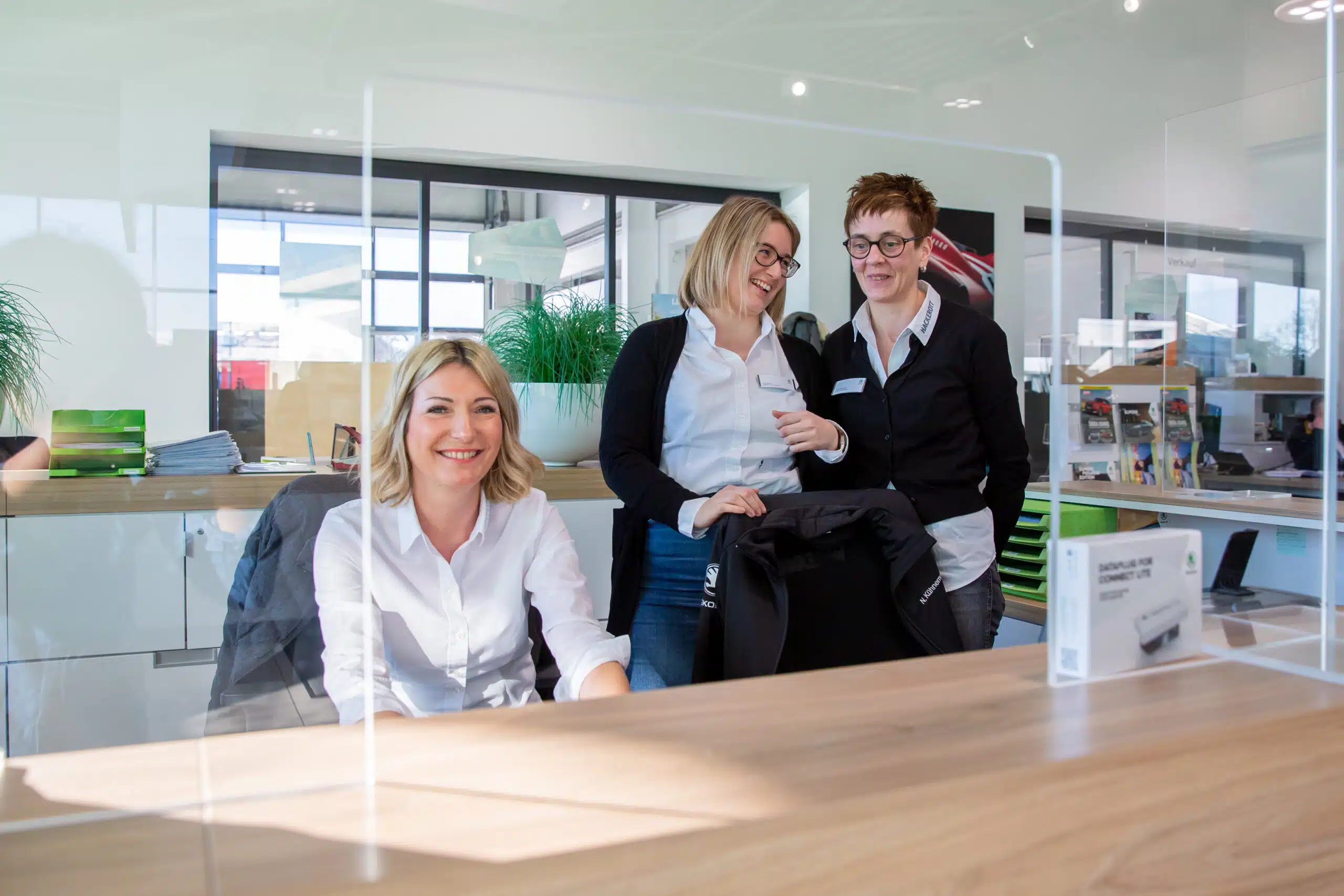 Drei lächelnde Hackerott-Mitarbeiterinnen hinter einem Empfangstresen mit Plexiglas-Schutz.